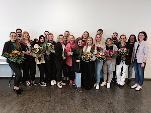Wundexperten 2026 Junge Menschen, teilweise mit Blumen in den Händen, stehen in mehreren Reihen nebeneinander vor einer weißen Wand und lächeln.